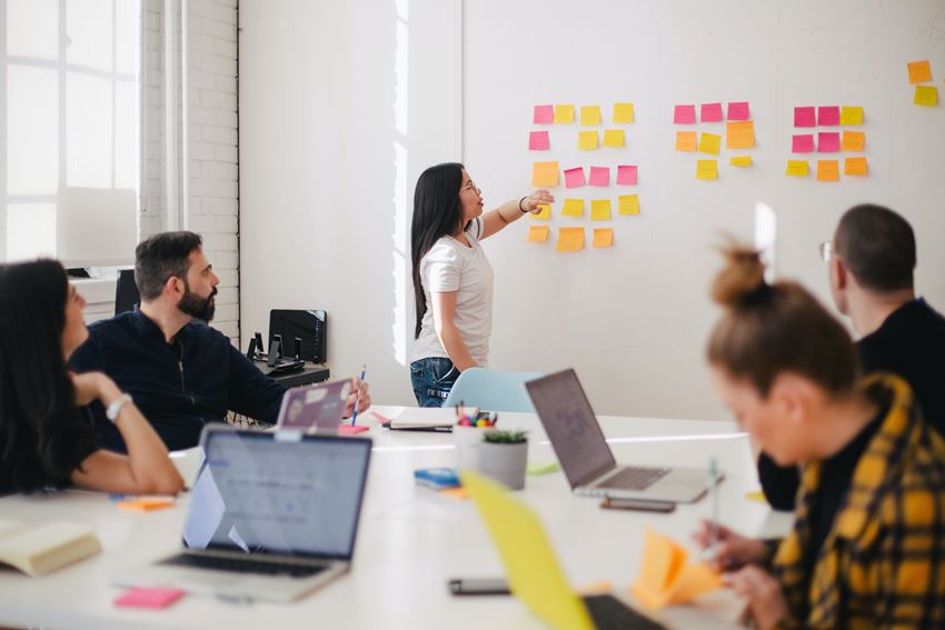 Flere personer sitter i et rom. En person står foran et whiteboard å peker på flere fargerike post it lapper som er hengt opp. Foto: Jason Goodman/Unsplash.