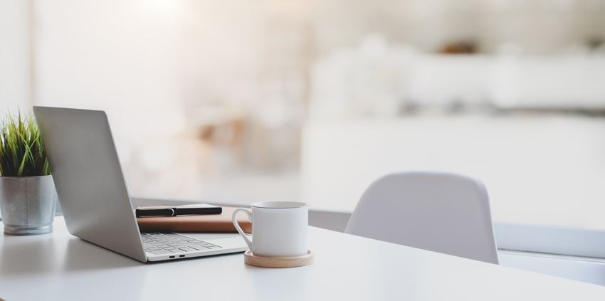 En grå pc, en hvit kaffekopp og notatbøker er på et skrivebord. Inntil bordet står det en grå stol.