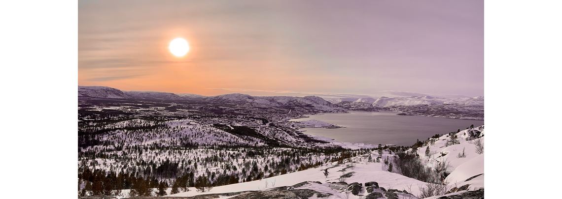 Landskapsbilde av snødekte fjell og skoger i solskinn.