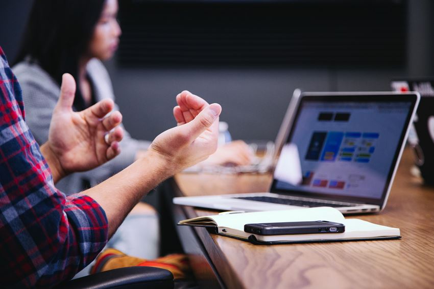 Bilde fokuserer på to armer foran en datamaskin, som har en notatbok og mobiltelefon foran seg. I bakgrunnen sitter et annen person å hører etter. Foto: Unsplash